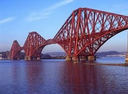 A large, red, steel cantilever bridge spans a body of water. The bridge has a complex latticework structure and multiple supporting piers. The water is relatively calm and reflects the bridge's structure. Land and some buildings are visible in the background on either side of the bridge, under a clear blue sky.
