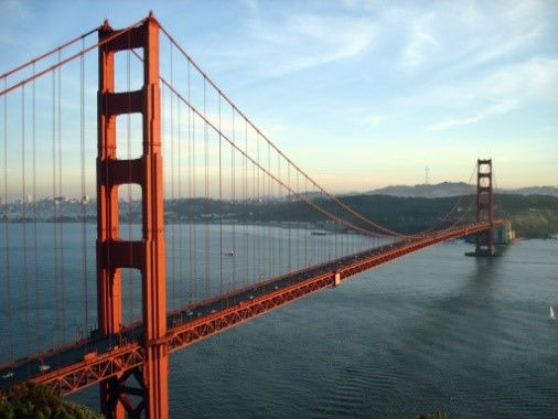 The Golden Gate Bridge, a red suspension bridge, spans a body of water. The perspective is from a high vantage point, showcasing the bridge's length and the surrounding landscape of hills and water. Vehicles can be seen travelling across the bridge. The sky is a mix of blue and white, suggesting daytime with some cloud cover. A small sailboat is visible in the lower right corner of the image.