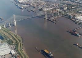 Aerial view of a cable-stayed bridge spanning a wide, muddy-looking river. Industrial buildings and port facilities are visible on both sides of the river, with ships and barges in the water.