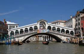 The Rialto Bridge in Venice, Italy, spans the Grand Canal. The white stone bridge, with its covered walkway lined with arches and shops, is crowded with people. Several gondolas and other boats navigate the canal beneath the bridge. Buildings line the canal on either side. The sky is clear and blue.