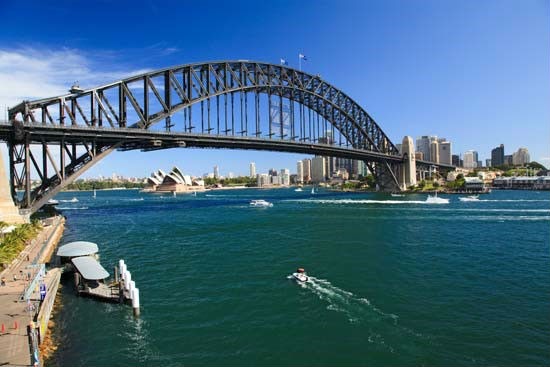 A steel arch bridge spans a body of dark teal water. The Sydney Opera House is visible in the middle distance to the left of the bridge. The Sydney city skyline is visible in the background on the right. Several boats are on the water. A pier with a covered structure is visible in the lower left foreground. The sky is clear and blue.
