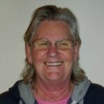 Chris Dann. A smiling lady with shoulder-length dark grey hair and glasses.