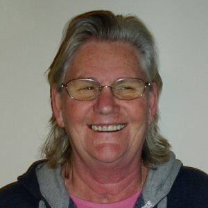 Chris Dann. A smiling lady with shoulder-length dark grey hair and glasses.